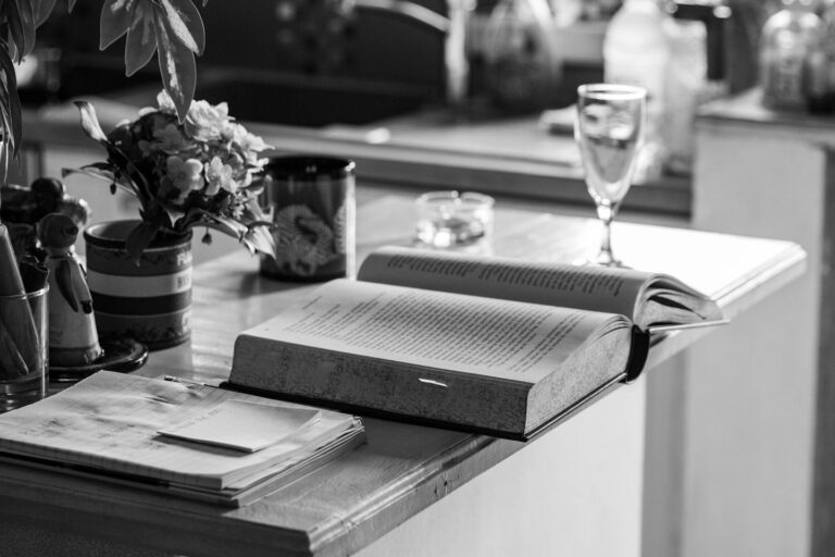 A black and white photo of an open book, with some flowers, a glass, and a notebook.